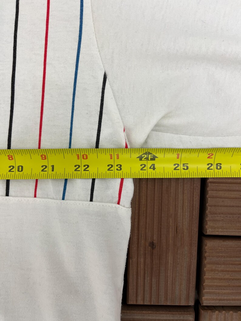 May include: A white shirt featuring vertical pinstripes in black, red, and blue. A yellow measuring tape is positioned across the fabric, showing a width measurement extending from 20 inches to 26 inches across the garment's midsection.