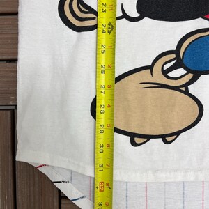 May include: A white t-shirt featuring a cartoon character graphic with black, red, blue, and tan colors. A yellow measuring tape is placed vertically over the shirt, showing a length measurement extending past 34 inches from the top of the graphic.