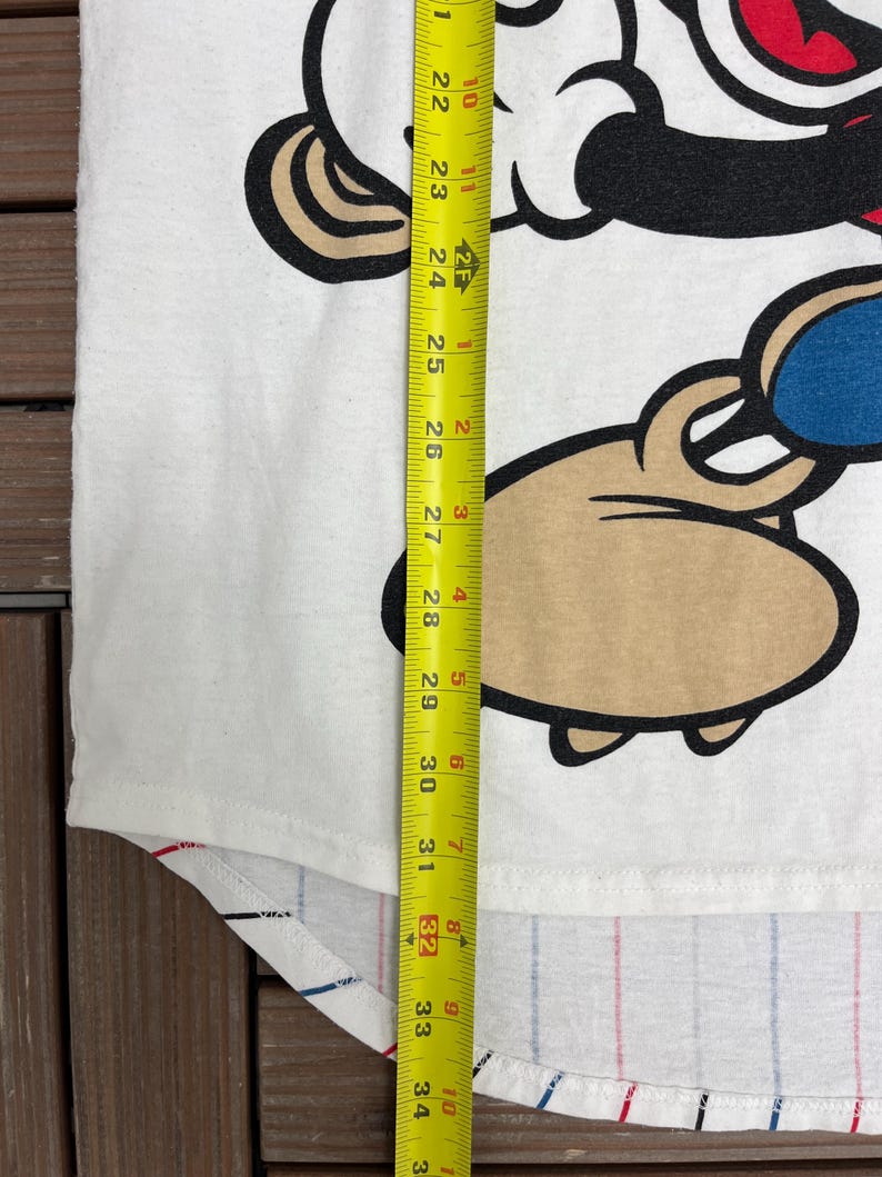 May include: A white t-shirt featuring a cartoon character graphic with black, red, blue, and tan colors. A yellow measuring tape is placed vertically over the shirt, showing a length measurement extending past 34 inches from the top of the graphic.