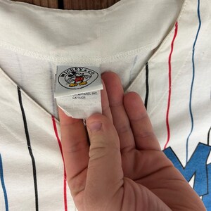 May include: A white baseball jersey featuring vertical pinstripes in red, blue, and black. The garment includes a white interior tag with the text MICKEY & CO, a graphic of Mickey Mouse, and the text APPAREL INC. CA 14426 printed below the logo.
