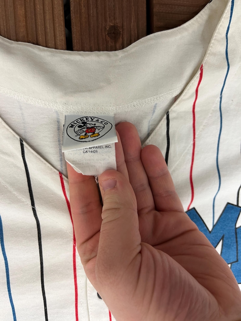 May include: A white baseball jersey featuring vertical pinstripes in red, blue, and black. The garment includes a white interior tag with the text MICKEY & CO, a graphic of Mickey Mouse, and the text APPAREL INC. CA 14426 printed below the logo.