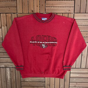 May include: A red long-sleeve sweatshirt featuring a black and white graphic on the chest that reads 49ERS SAN FRANCISCO with a team logo. The garment has a ribbed crew neckline and cuffs with dark grey stripes and a Starter brand label inside.