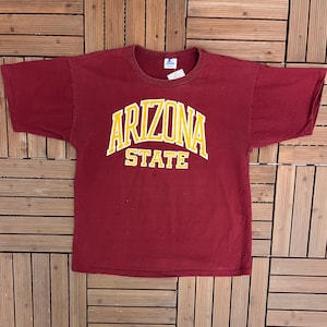 Peut inclure: T-shirt bordeaux à manches courtes avec l'inscription "ARIZONA STATE" en lettres jaunes et blanches sur la poitrine. Le t-shirt est posé à plat sur une surface en bois.