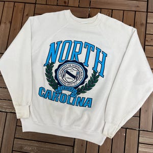 K&ouml;nnte beinhalten: Ein wei&szlig;es Sweatshirt mit dem Schriftzug "NORTH CAROLINA" in blauer Schrift. Das Wort "NORTH" ist oben und "CAROLINA" unten gebogen. In der Mitte befindet sich ein kreisf&ouml;rmiges Emblem mit Text und einem Lorbeerkranz.