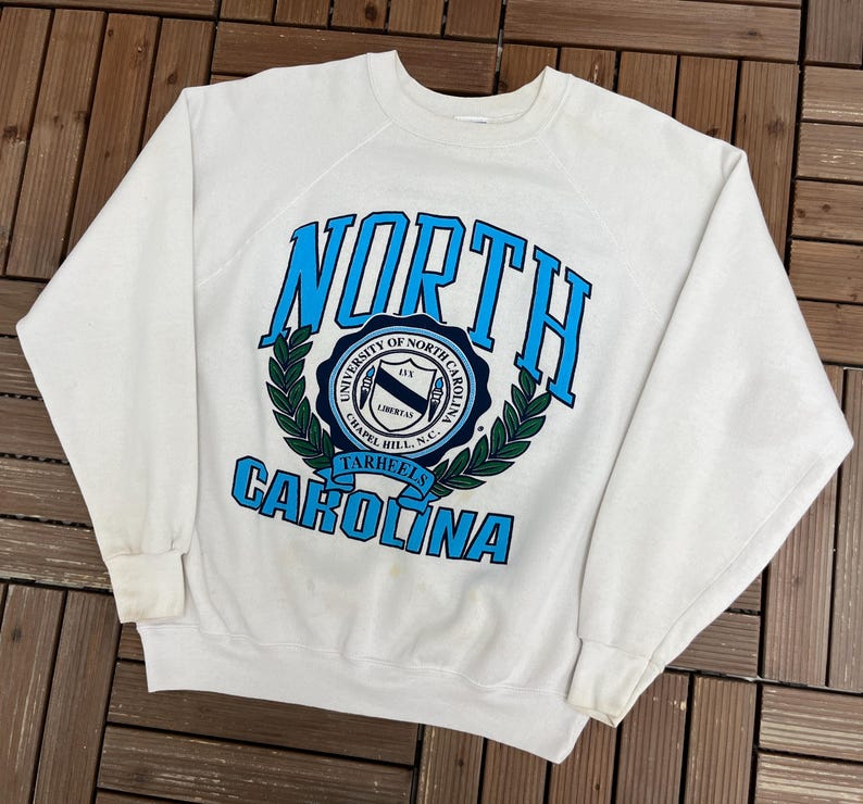 K&ouml;nnte beinhalten: Ein wei&szlig;es Sweatshirt mit dem Schriftzug "NORTH CAROLINA" in blauer Schrift. Das Wort "NORTH" ist oben und "CAROLINA" unten gebogen. In der Mitte befindet sich ein kreisf&ouml;rmiges Emblem mit Text und einem Lorbeerkranz.