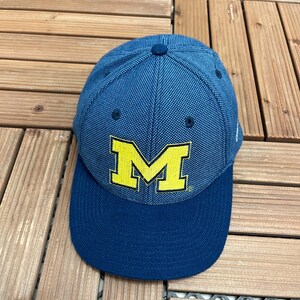 May include: A blue baseball cap with a textured fabric crown and a solid dark blue brim. The front of the cap features a large, embroidered yellow letter M with a thin black outline. The cap has a button on top and small ventilation eyelets.