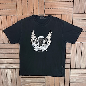 May include: Black t-shirt featuring a white and gray graphic of the Guinness logo with wings and a banner that reads "Brewed in Ireland" and "Guinness ESTD 1759". The shirt is made of a soft material.