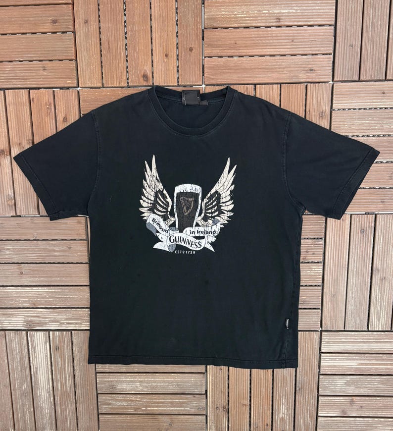 May include: Black t-shirt featuring a white and gray graphic of the Guinness logo with wings and a banner that reads "Brewed in Ireland" and "Guinness ESTD 1759". The shirt is made of a soft material.