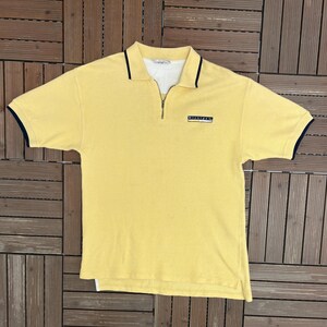 Peut inclure: Polo jaune avec col zippé, bordure bleu marine sur le col et les manches, et le mot "Michigan" sur la poitrine. Le t-shirt est fait d'un tissu doux et texturé.