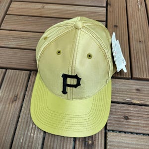 May include: A gold baseball cap featuring a black "P" logo. The cap has a curved peak and a structured crown. The cap is made of a durable fabric and has a tag attached.