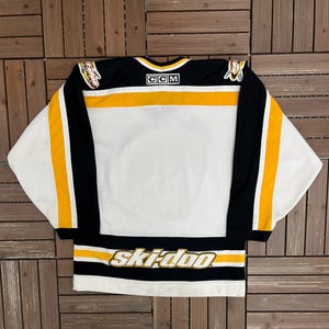 May include: White hockey jersey with black and gold accents. The jersey features the text "ski-doo" across the bottom and the CCM logo on the collar. The sleeves have black and gold stripes.