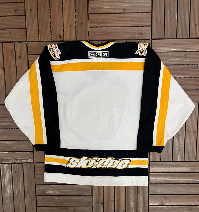 May include: White hockey jersey with black and gold accents. The jersey features the text "ski-doo" across the bottom and the CCM logo on the collar. The sleeves have black and gold stripes.