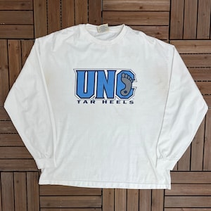 Könnte beinhalten: Ein weißes Langarm-T-Shirt mit dem blauen Text "UNC TAR HEELS". Das Design beinhaltet die Buchstaben "UNC" und eine Fußgrafik. Das Shirt besteht aus einem weichen Material und hat einen Rundhalsausschnitt.