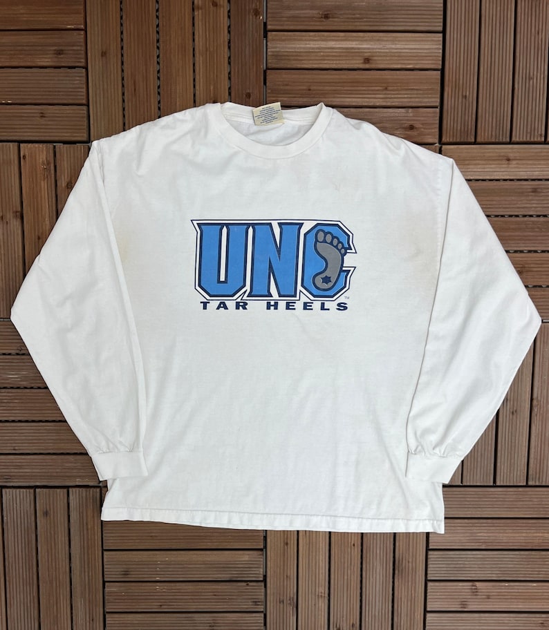 May include: A white long-sleeved t-shirt with the blue text "UNC TAR HEELS". The design includes the letters "UNC" and a foot graphic. The shirt is made of a soft material and has a crew neck.