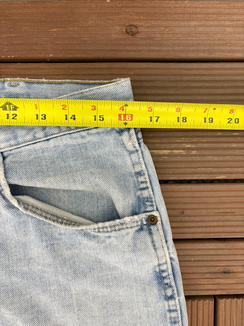 May include: Light blue denim jeans with a pocket and a yellow measuring tape showing a measurement of 16 inches. The jeans are on a wooden surface.