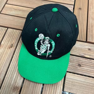 May include: Black and green baseball cap with the Boston Celtics logo. The cap has a green peak and a black crown with green details. The logo shows a leprechaun with a basketball. A green button is on top.