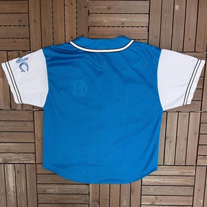 May include: A blue baseball jersey with white sleeves. The jersey has a white collar and black stripes on the sleeves. The letters "NC" are visible on the left sleeve. The jersey is made of a mesh material.