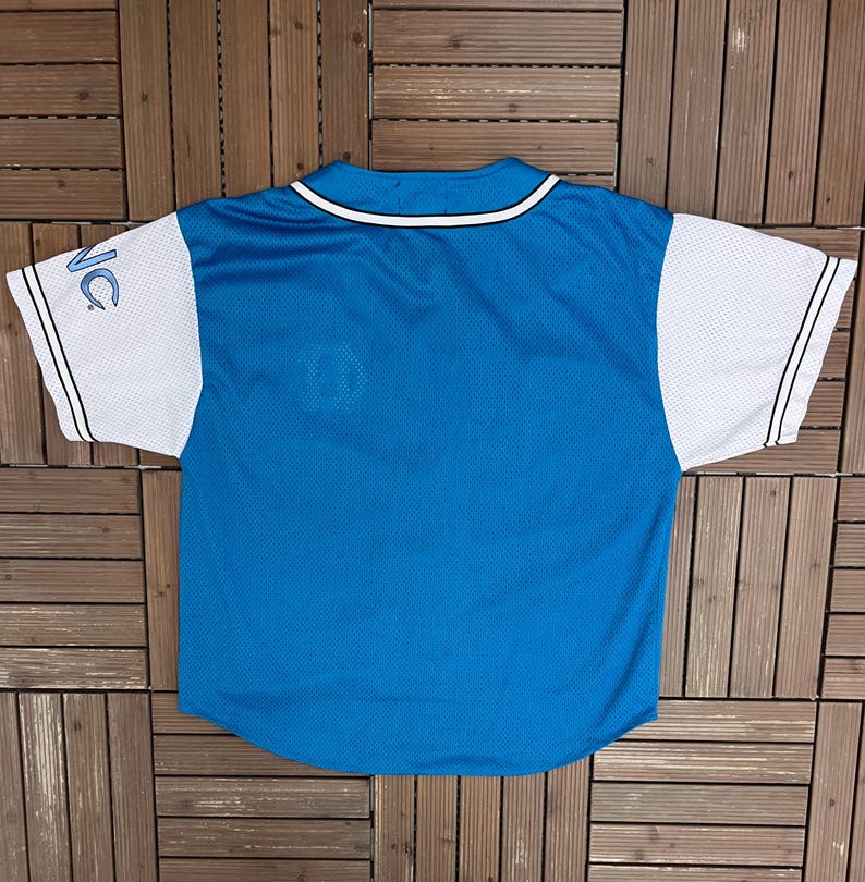 May include: A blue baseball jersey with white sleeves. The jersey has a white collar and black stripes on the sleeves. The letters "NC" are visible on the left sleeve. The jersey is made of a mesh material.