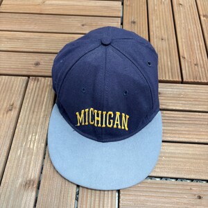 May include: A navy blue baseball cap with a grey peak. The word "MICHIGAN" is embroidered in gold across the front. The cap is on a wooden surface.