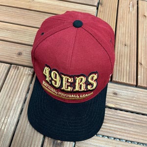 Pode incluir: Boné de beisebol bordô com aba preta. O boné apresenta o texto "49ERS" em letras douradas, com "NATIONAL FOOTBALL LEAGUE" abaixo. Possui um botão preto na parte superior.