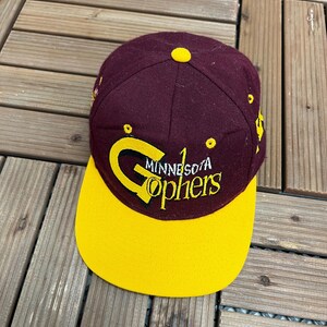 Puede incluir: Gorra de béisbol granate y dorada con la inscripción "Minnesota Gophers" bordada en dorado. La gorra tiene una visera amarilla y un botón amarillo. Fabricada en tejido resistente.