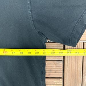 May include: A black t-shirt with a yellow measuring tape laid across the front. The tape measures approximately 9 inches across the chest. The shirt's sleeve is visible on the right side of the image.