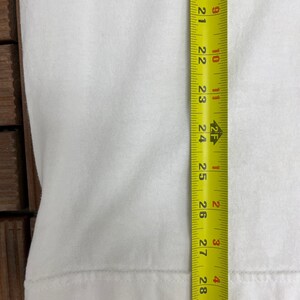 May include: A white t-shirt is shown with a yellow measuring tape next to it. The tape measures from 50.8 cm to 73.7 cm. The shirt is plain and has a simple hem.