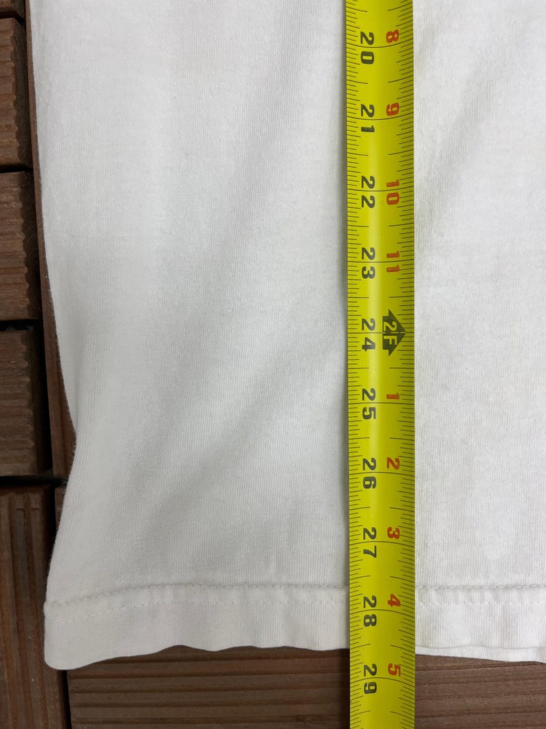 May include: A white t-shirt is shown with a yellow measuring tape next to it. The tape measures from 50.8 cm to 73.7 cm. The shirt is plain and has a simple hem.
