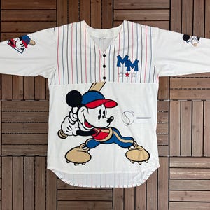May include: A white baseball jersey with red and blue pinstripes, featuring a large graphic of Mickey Mouse holding a baseball bat. The shirt has three buttons, the letters MM in blue, and small baseball-themed graphics on the sleeves.