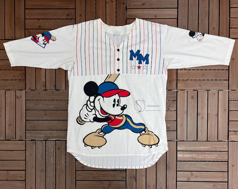 May include: A white baseball jersey with red and blue pinstripes, featuring a large graphic of Mickey Mouse holding a baseball bat. The shirt has three buttons, the letters MM in blue, and small baseball-themed graphics on the sleeves.
