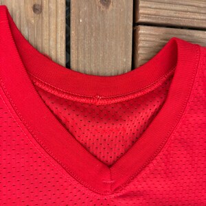 May include: Close-up of a bright red V-neck jersey. The jersey has a mesh fabric with small holes throughout. The neckline is reinforced with a solid red band. The jersey is likely for sports or athletic wear.