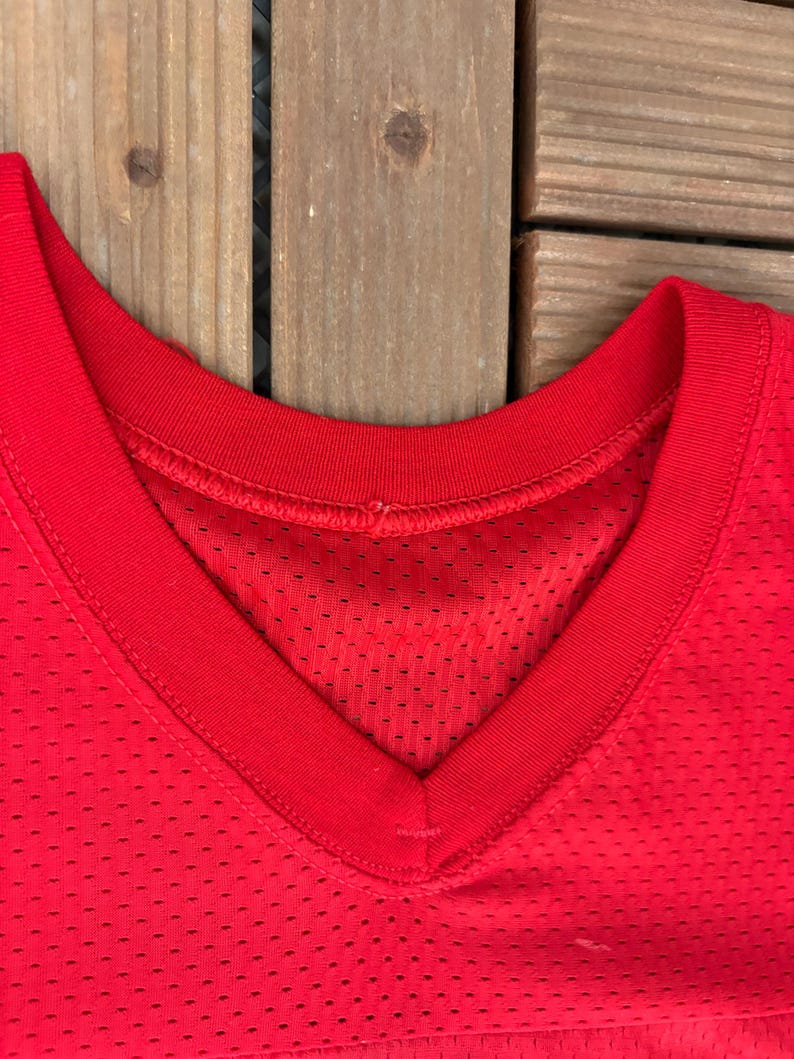 May include: Close-up of a bright red V-neck jersey. The jersey has a mesh fabric with small holes throughout. The neckline is reinforced with a solid red band. The jersey is likely for sports or athletic wear.