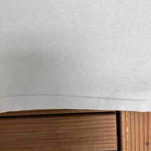 May include: Close-up of a light blue t-shirt. The fabric is smooth and appears to be a knit material. The bottom hem is visible, with a line of stitching. The shirt is resting on a wooden surface.