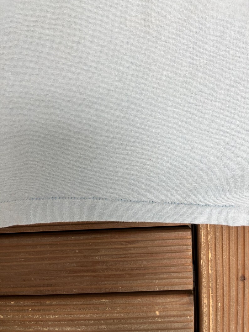 May include: Close-up of a light blue t-shirt. The fabric is smooth and appears to be a knit material. The bottom hem is visible, with a line of stitching. The shirt is resting on a wooden surface.