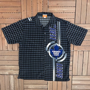 May include: Black and grey button-down shirt with a maple leaf pattern. Features the Toronto Maple Leafs logo and the word "LEAFS" in blue and white. Short-sleeved, with a collared neckline.