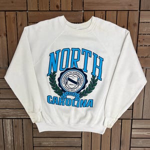 Könnte beinhalten: Ein weißes Sweatshirt mit Rundhalsausschnitt und dem Schriftzug "NORTH CAROLINA" in blauer Schrift. Das Sweatshirt hat ein rundes Emblem mit Text und einem Lorbeerkranz-Design. Der Text "UNIVERSITY OF NORTH CAROLINA CHAPEL HILL, N.C. TARHEELS" ist ebenfalls sichtbar.