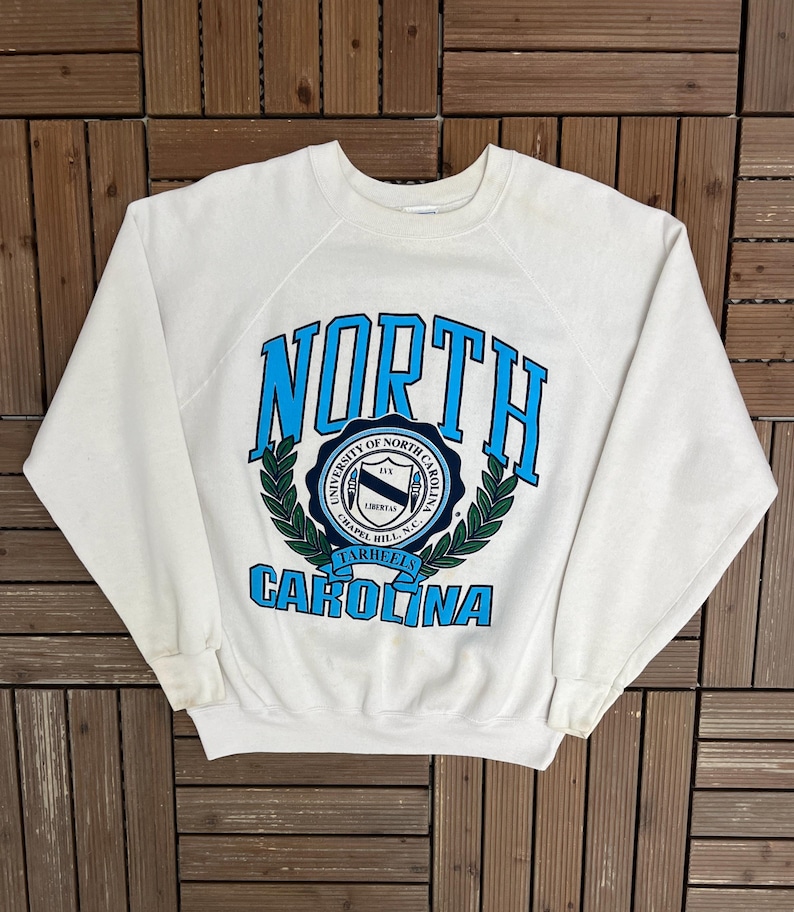 K&ouml;nnte beinhalten: Ein wei&szlig;es Sweatshirt mit Rundhalsausschnitt und dem Schriftzug "NORTH CAROLINA" in blauer Schrift. Das Sweatshirt hat ein rundes Emblem mit Text und einem Lorbeerkranz-Design. Der Text "UNIVERSITY OF NORTH CAROLINA CHAPEL HILL, N.C. TARHEELS" ist ebenfalls sichtbar.