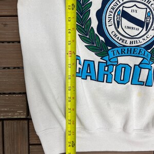 K&ouml;nnte beinhalten: Wei&szlig;es Sweatshirt mit den Worten "NORTH CAROLINA" in hellblauen Buchstaben. Darunter befindet sich ein marineblaues Siegel mit den Worten "UNIVERSITY OF NORTH CAROLINA CHAPEL HILL, N.C." und dem Wort "TARHEELS". Das Sweatshirt ist ca. 66 cm lang.