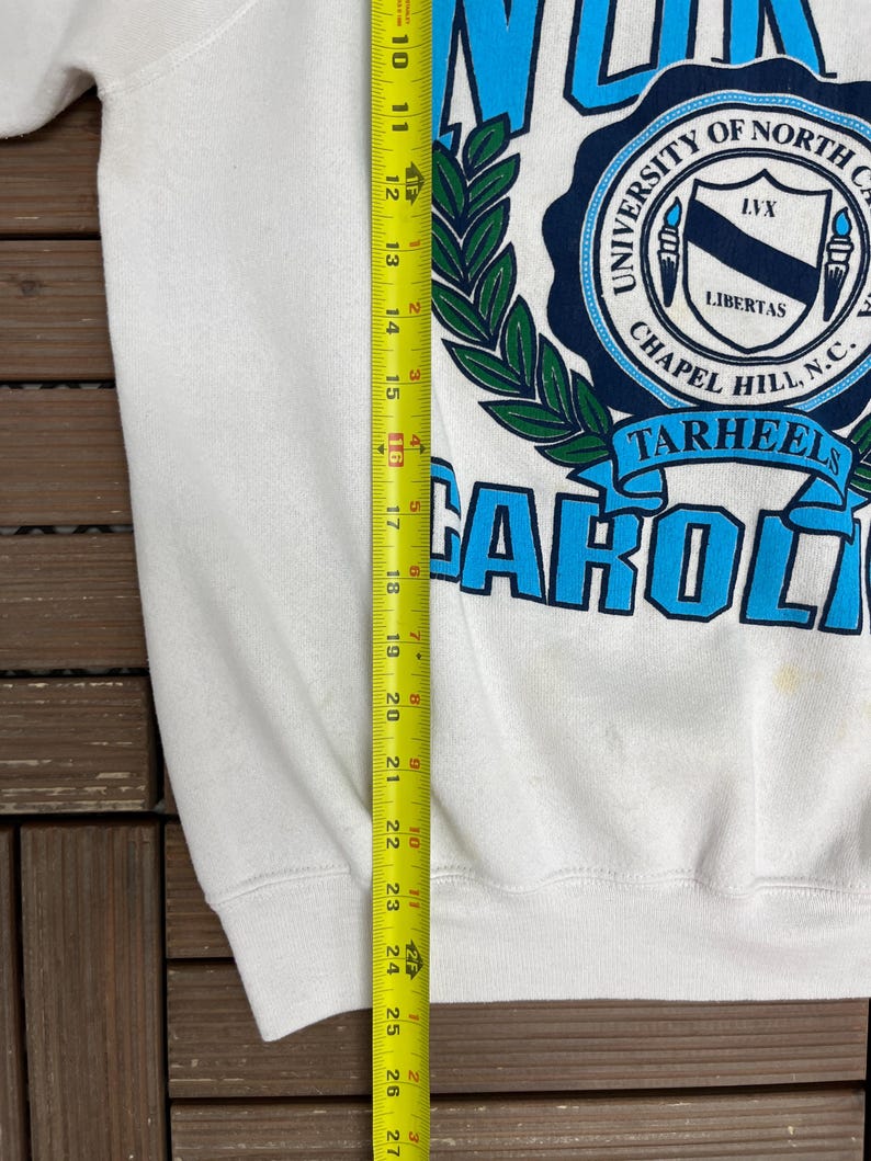 K&ouml;nnte beinhalten: Wei&szlig;es Sweatshirt mit den Worten "NORTH CAROLINA" in hellblauen Buchstaben. Darunter befindet sich ein marineblaues Siegel mit den Worten "UNIVERSITY OF NORTH CAROLINA CHAPEL HILL, N.C." und dem Wort "TARHEELS". Das Sweatshirt ist ca. 66 cm lang.