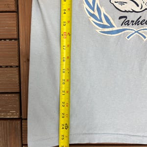 May include: Light blue t-shirt with a graphic of a cartoon frog wearing a crown and the word "Tarhees". The shirt is measured with a tape measure, showing a length of approximately 27 inches.