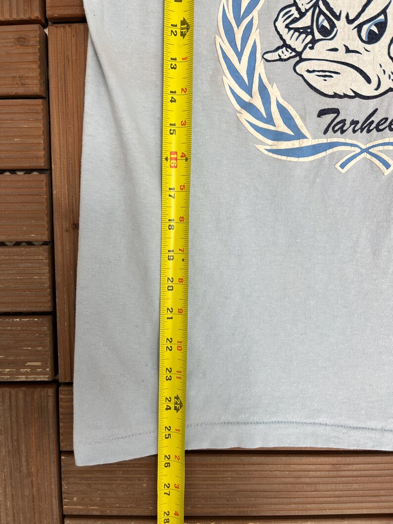 May include: Light blue t-shirt with a graphic of a cartoon frog wearing a crown and the word "Tarhees". The shirt is measured with a tape measure, showing a length of approximately 27 inches.