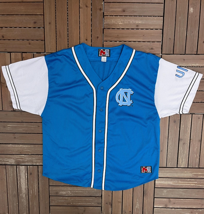 May include: A light blue baseball jersey with white sleeves. The jersey features a button-up front, a V-neck, and the logo of the University of North Carolina on the chest. The sleeves have the word "UN" on them.