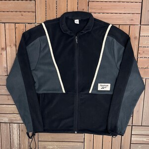 Peut inclure: Veste zippée noire avec des panneaux gris sur les manches et les épaules. La veste présente un motif à rayures blanches et le logo Reebok sur le devant. La veste a un cordon de serrage en bas.
