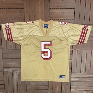 May include: A gold-coloured athletic jersey featuring the number 5 in white with a red outline on the chest. The jersey has a V-neck collar, short sleeves with red and black stripes, and the Adidas logo on the sleeve and a tag at the bottom hem.