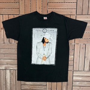 May include: Black t-shirt featuring a graphic of Terri Clark in a white suit and hat. The image is framed with a decorative border. The text "TERRI CLARK" is printed at the top of the graphic.