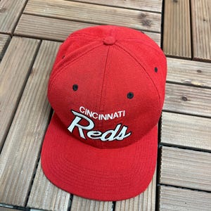 Może przedstawiać: Czerwona czapka bejsbolowa z napisem "CINCINNATI Reds" w białym skrypcie. Czapka ma zakrzywiony daszek i czarne oczka. Wykonana z wytrzymałej tkaniny.