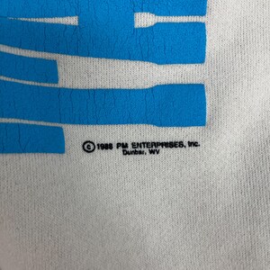 K&ouml;nnte beinhalten: Nahaufnahme eines wei&szlig;en Stoffes mit gro&szlig;en, blauen, horizontalen Streifen. Der Text "1988 PM ENTERPRISES, Inc. Dunbar, WV" ist in Schwarz gedruckt. Der Stoff scheint ein T-Shirt oder Sweatshirt zu sein.
