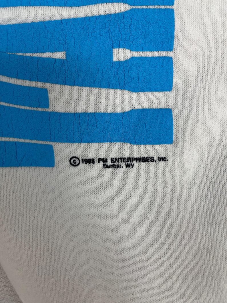 K&ouml;nnte beinhalten: Nahaufnahme eines wei&szlig;en Stoffes mit gro&szlig;en, blauen, horizontalen Streifen. Der Text "1988 PM ENTERPRISES, Inc. Dunbar, WV" ist in Schwarz gedruckt. Der Stoff scheint ein T-Shirt oder Sweatshirt zu sein.