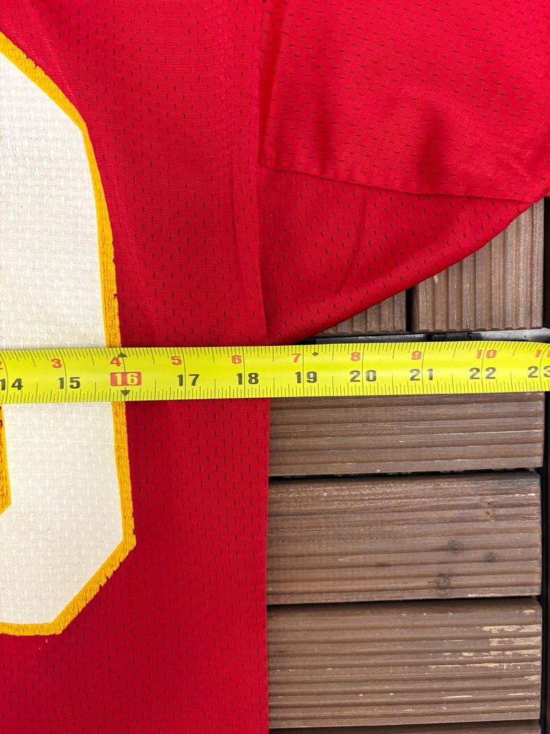May include: Red mesh jersey with a white and yellow number. A yellow measuring tape shows the jersey is approximately 40.6 cm wide. The jersey is on a wooden surface.
