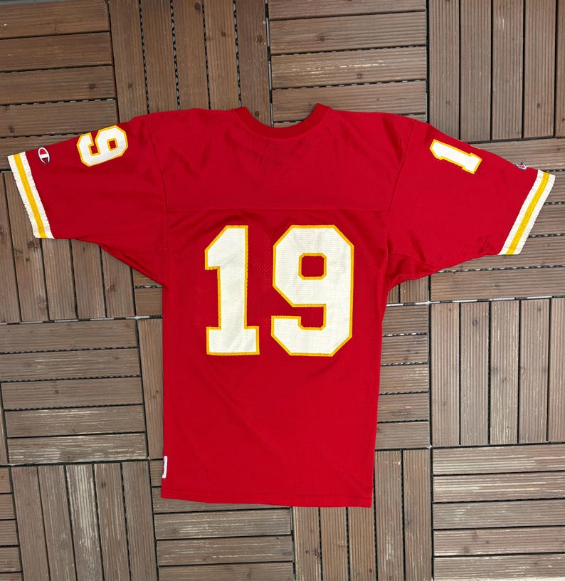 May include: A red Champion jersey featuring the number 19 in white with a gold outline on the front. The sleeves display the numbers 1 and 9 in white with a gold outline. The jersey has gold and white stripes on the sleeves.